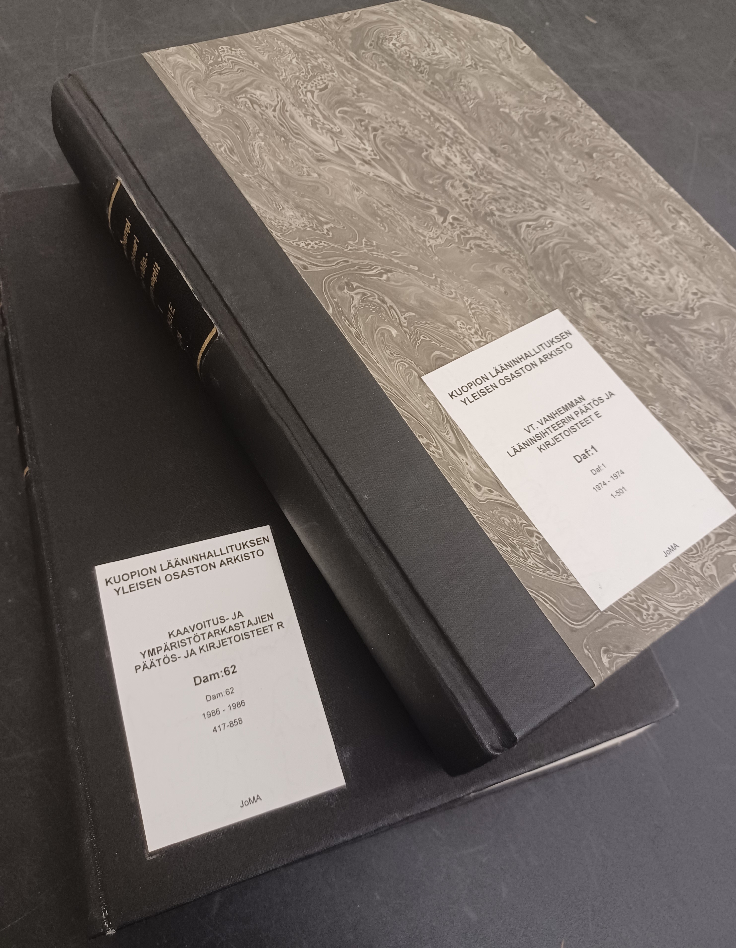 Pöydällä on kaksi suljettua kirjaa, joiden etukannessa on nimiö ja siinä arkistoyksikön tiedot.